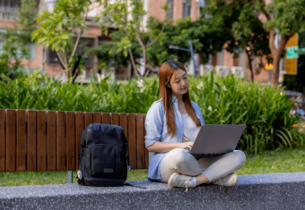 A student sitting on the ground next to their backpack looking for jobs on their laptop.